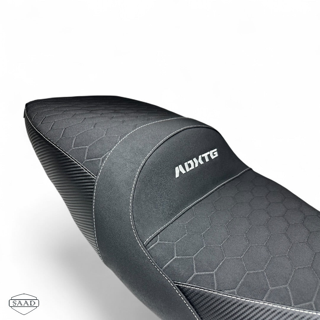 Close-up of a black motorcycle comfort seat with a carbon fiber weave pattern and a honeycomb texture, featuring the brand logo 'ADXTG' on the side